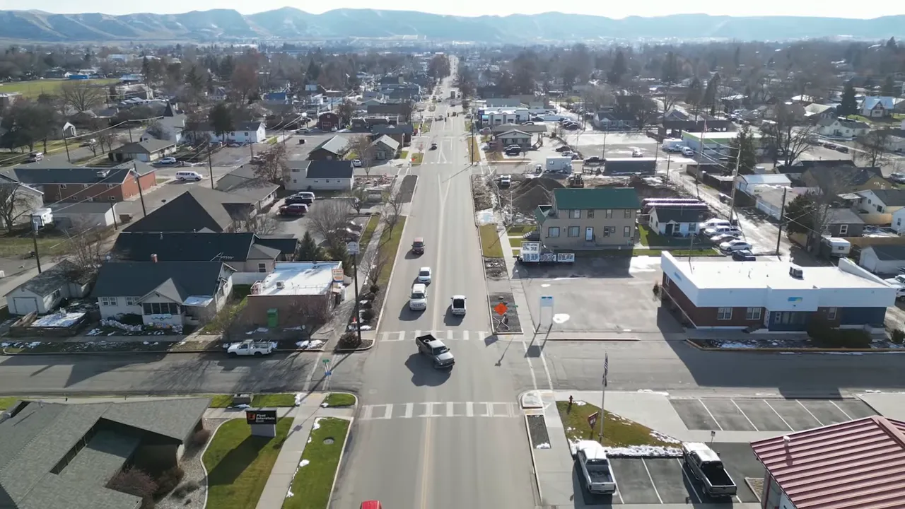 Street-level aerial view down a small town main street with shops, parked cars and a church steeple