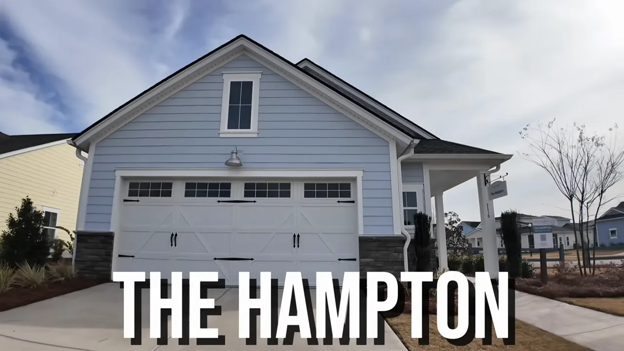 Host carrying a child while following the presenter down the Hampton model’s long covered porch with light-blue hardy plank siding, stone knee wall and the front door at the end.