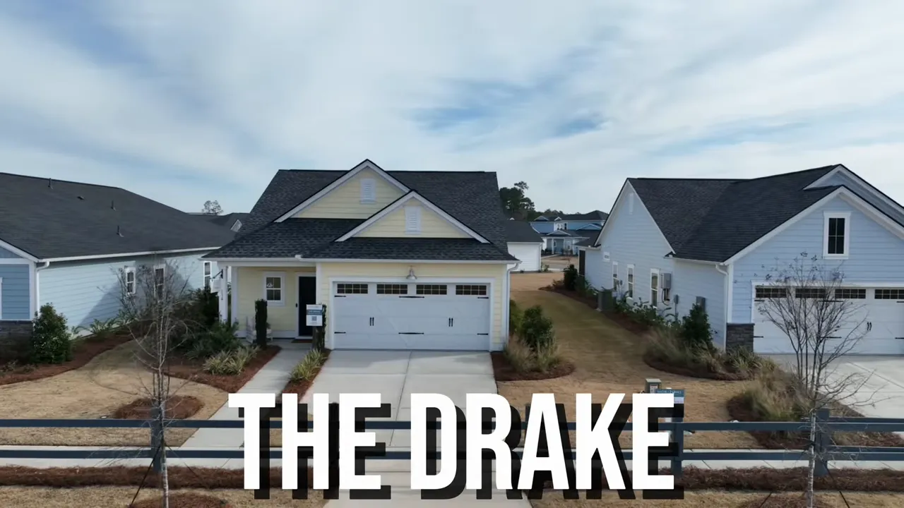 Presenter at the front entry of The Drake model home with a 'THE DRAKE' sign visible by the doorway and the model's yellow siding.