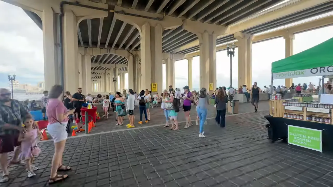 Vendor and interviewer under a red market tent at a Jacksonville arts market with colorful display and signage