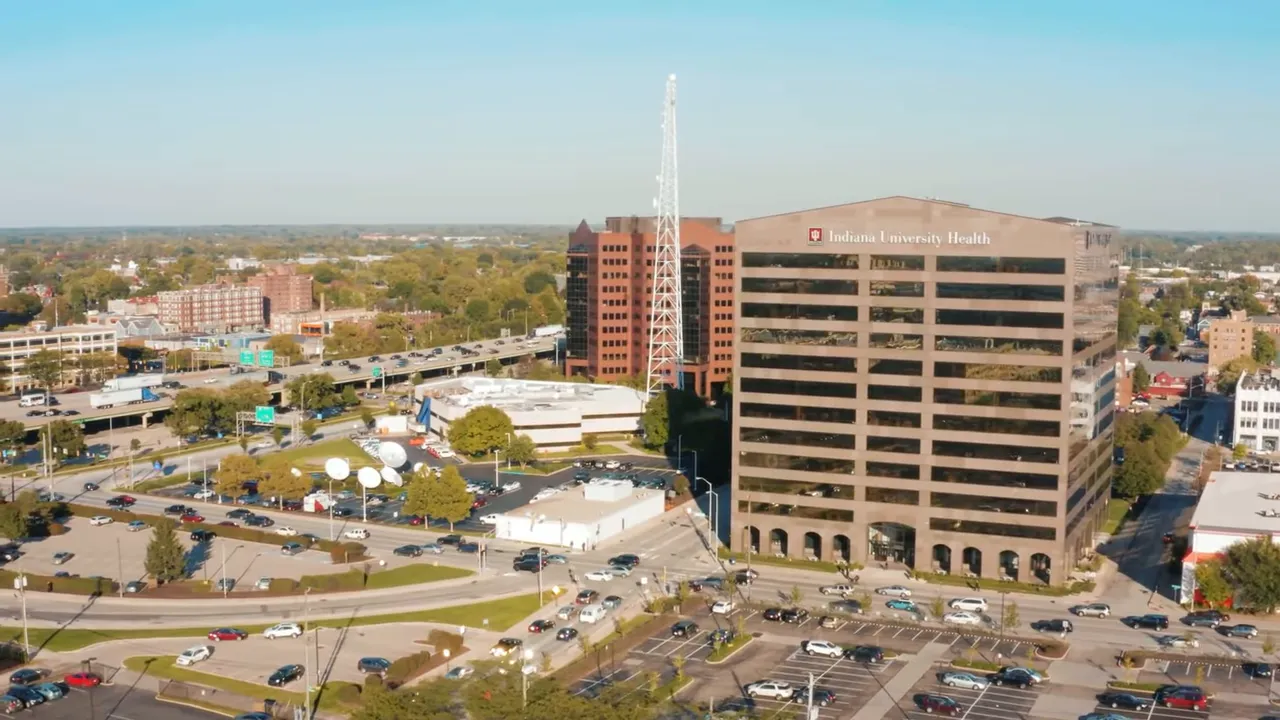 Wide shot of the host in front of Indianapolis buildings and skyline with bright green text overlay 'ADDS $7B+ OF INVESTMENT'.