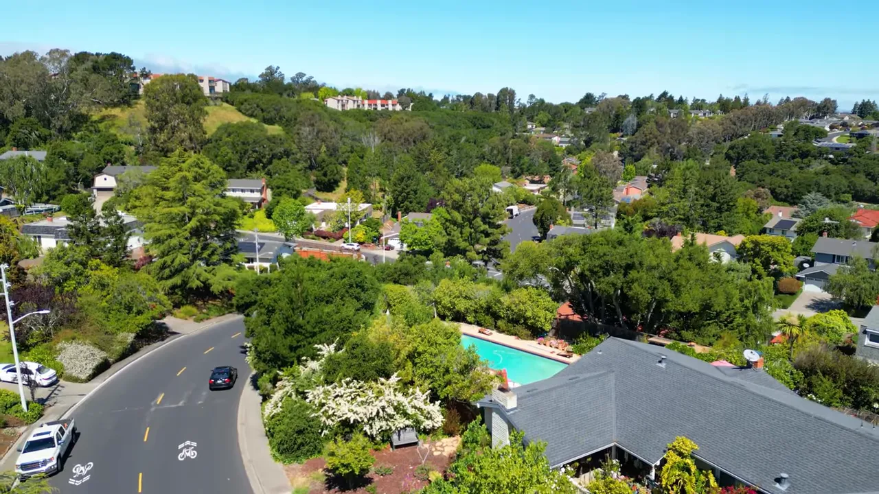 Presenter standing on a sidewalk in front of a house with deep shrubbery and raised lot, showing space and privacy on a hillside street