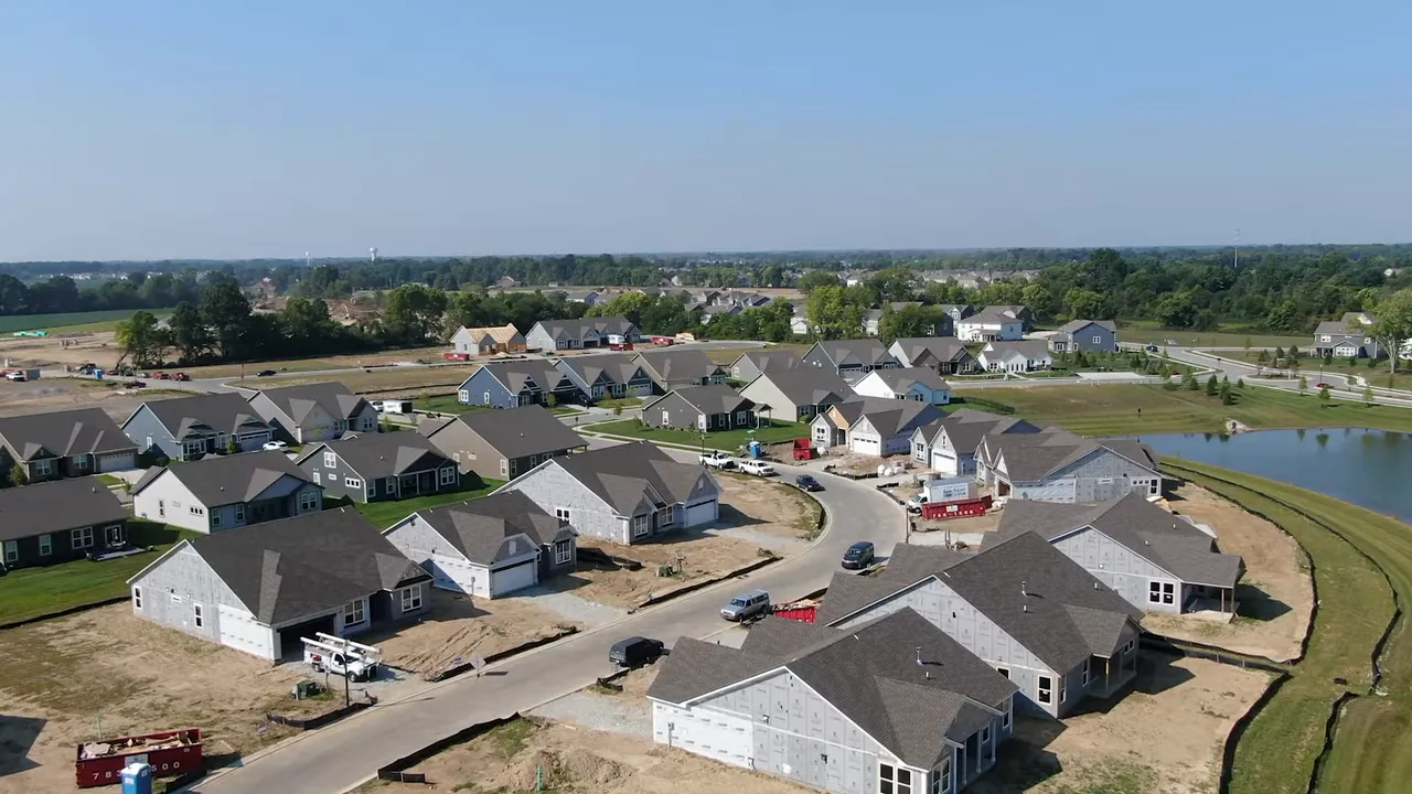 Presenter speaking on a downtown sidewalk in front of a mixed‑use building and a historical marker, clear framing and hand gesture