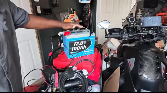 Blue 12.8V 100Ah battery in a garage beside motorcycles and a coiled air compressor hose