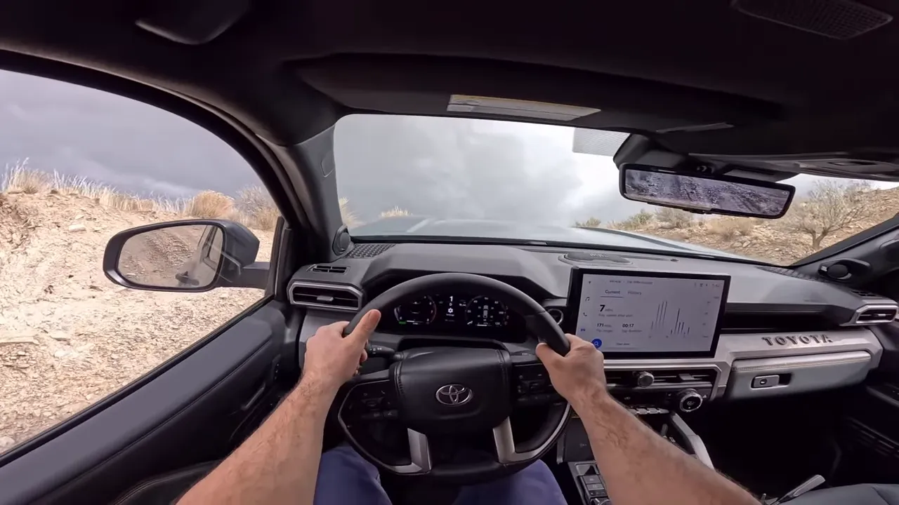 Centered interior view from the driver's seat of a Toyota 4Runner Trailhunter driving up a steep dirt hill, showing the hood, steering wheel and surrounding terrain.