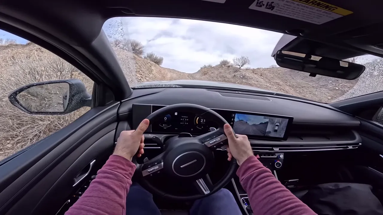 Driver POV showing steering wheel, digital instrument cluster and 360-degree camera display while navigating a rocky trail