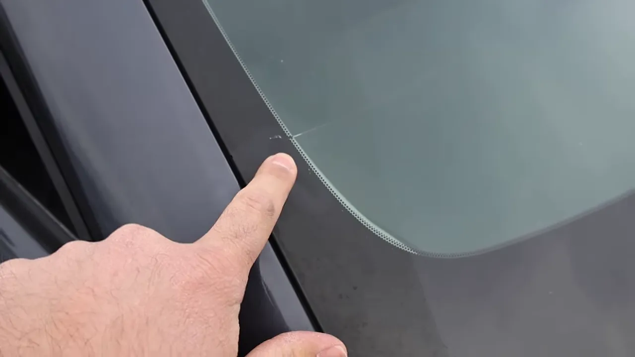 Finger pointing at a visible crack that starts at the black ceramic frit along the edge of a CX-50 windshield, with frit dots visible.