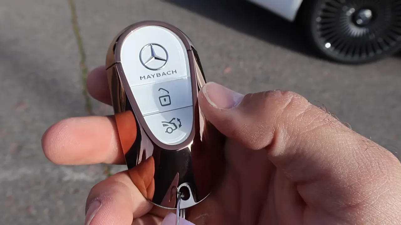 Hand holding Maybach key fob with Mercedes emblem and trunk button, car wheel visible in background