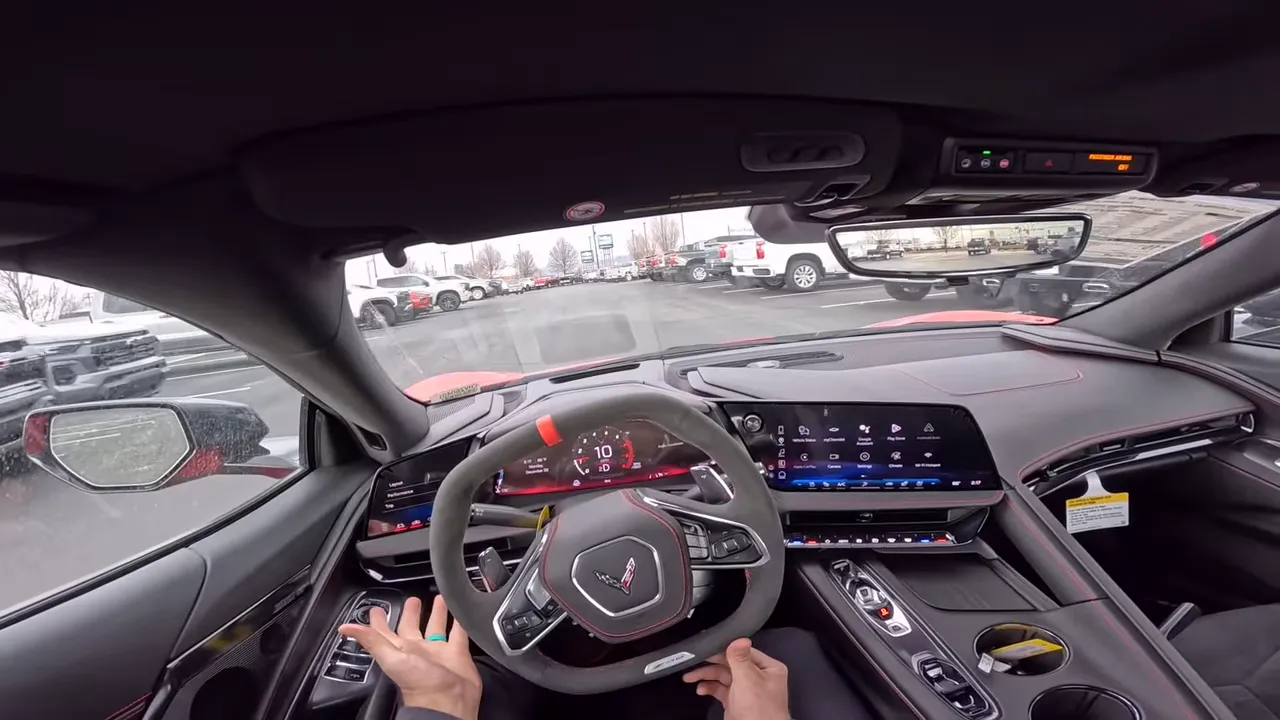 Centered driver point‑of‑view inside a 2026 Corvette Z06 with steering wheel, digital gauges and a clear view of dealership trucks and cars through the windshield and rearview mirror.