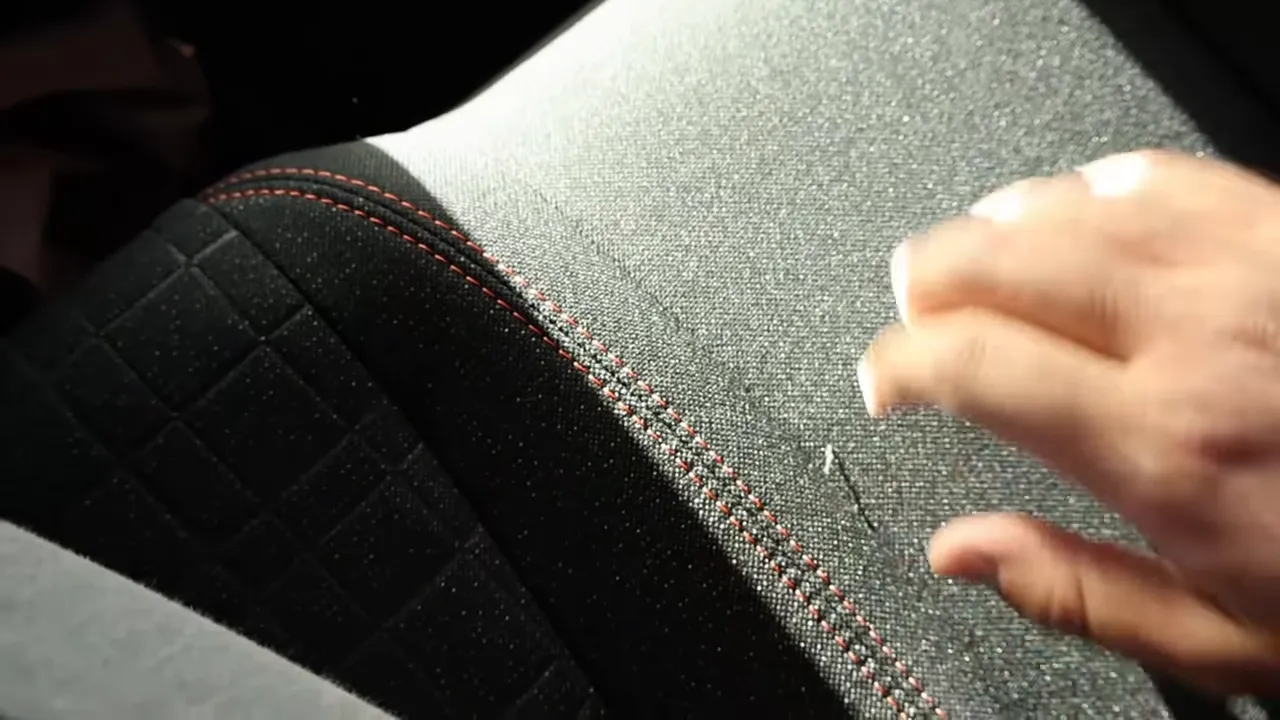 Close‑up of a seat showing contrast red/orange stitching and cloth upholstery with a hand indicating material quality.