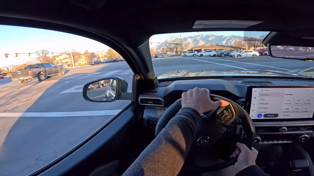 Driver POV inside a Toyota 4Runner Trailhunter approaching an intersection with a dealership lot visible on the right, multiple parked cars and mountain backdrop