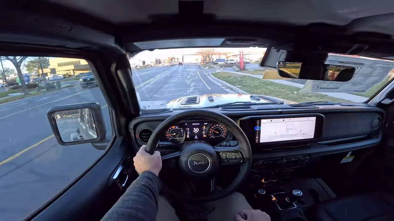 Centered driver POV inside a 2026 Jeep Wrangler Rubicon showing steering wheel, clear digital instrument cluster, infotainment screen and a straight road ahead.