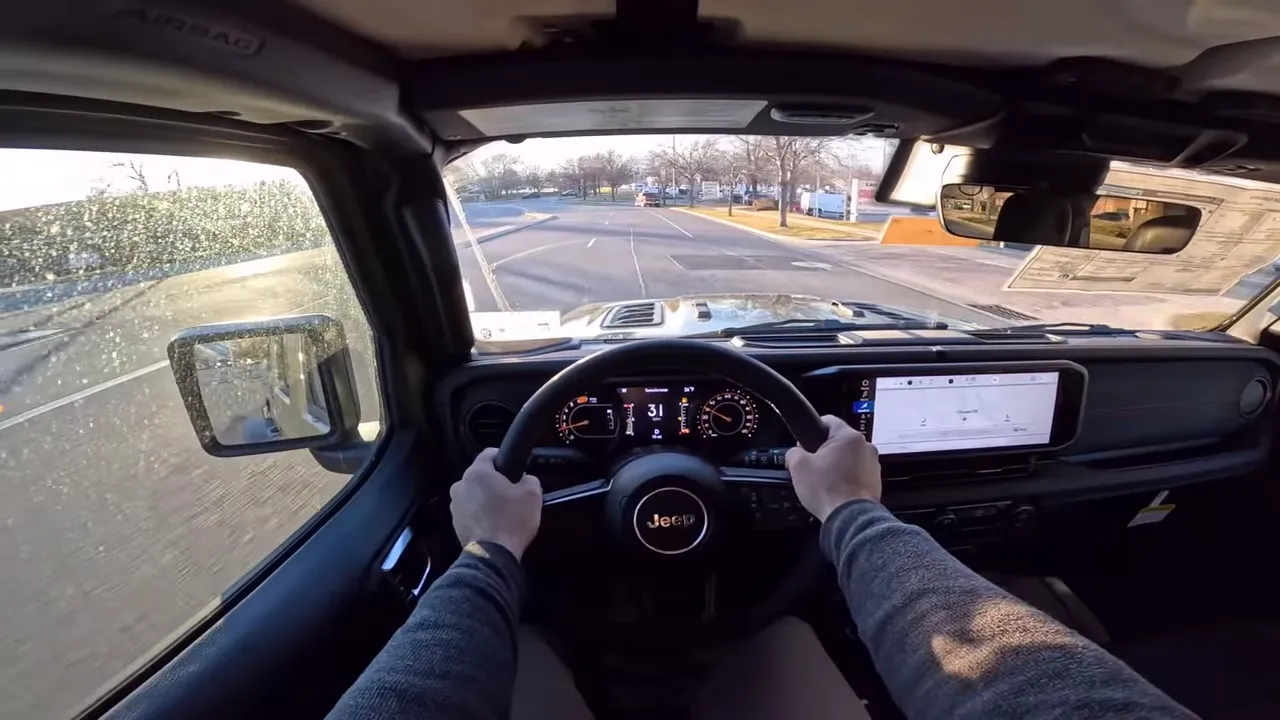 Centered driver POV inside a 2026 Jeep Wrangler showing steering wheel, clear digital gauges, infotainment screen and the road ahead