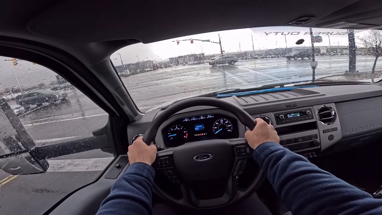 Driver's perspective inside a Ford F-650 showing steering wheel, instrument cluster, and rain‑dotted windshield at an intersection.