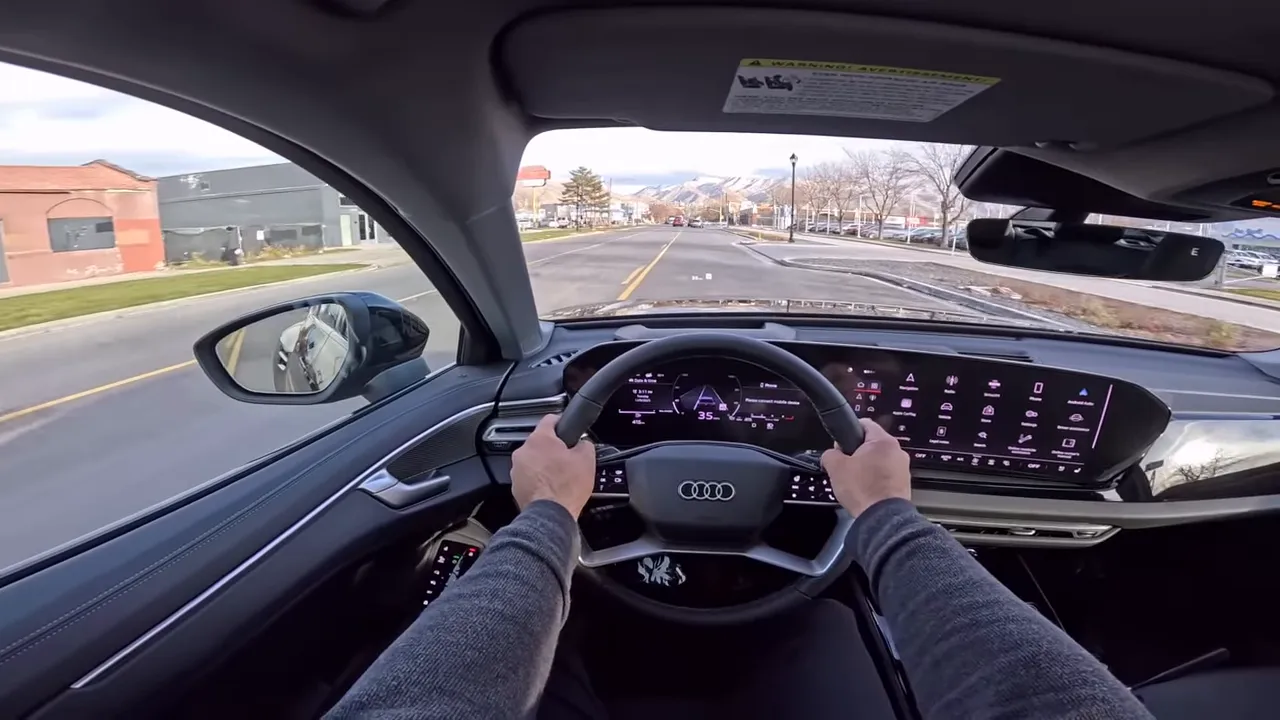 Driver point-of-view inside 2026 Audi A6 with both hands on steering wheel, digital instrument cluster, infotainment screen and road ahead visible.
