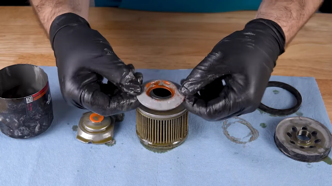 Gloved hands removing the top plate from an oil filter element revealing an orange gasket and contaminant residue around the opening