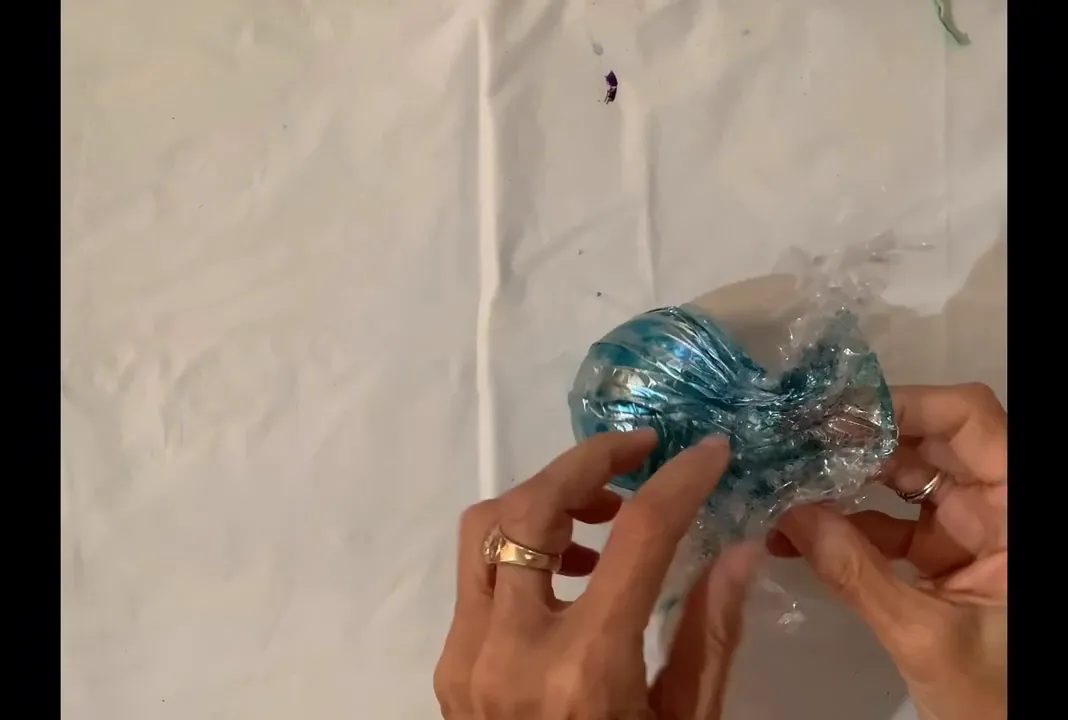 Hands unwrapping plastic-wrapped glass ornament showing blue alcohol-ink marbling and metallic highlights
