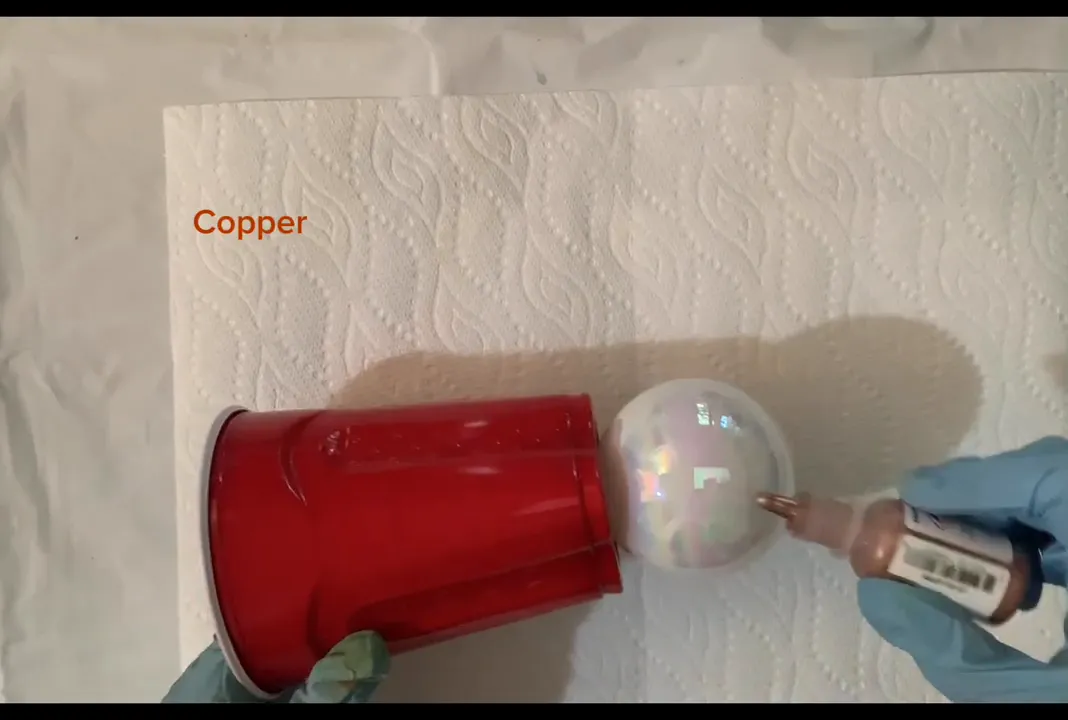 Applying copper alcohol ink to a white pearlescent glass ornament held in a red plastic cup on a paper towel