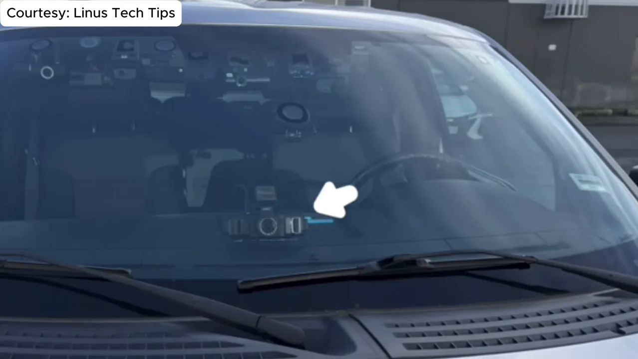 Close-up of a dash cam mounted in the center of the windshield behind the rearview mirror with a small arrow pointing to the unit.