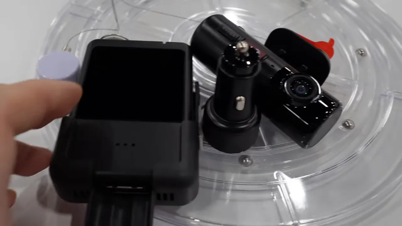 Clear overhead shot of a compact cylindrical dash camera with an SOS button, a cigarette-adapter power plug and a small touchscreen/display housing on a transparent exhibit stand.