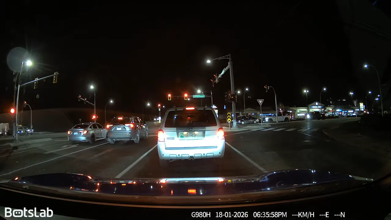 Nighttime dash cam view stopped behind a white SUV at a red light with plate area overexposed
