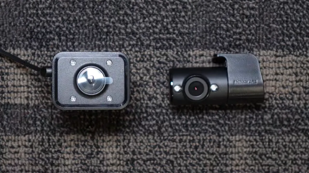 Two small interior dash cameras side-by-side on carpet — square IR camera with four LEDs (left) and cylindrical Thinkware-style camera (right).