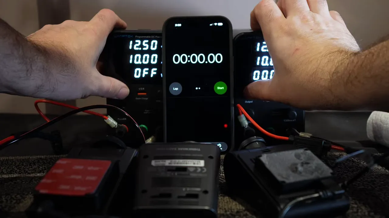 Dash cam test bench with smartphone stopwatch showing 00:00.00, two power supplies and three dash cameras ready for simultaneous power-on