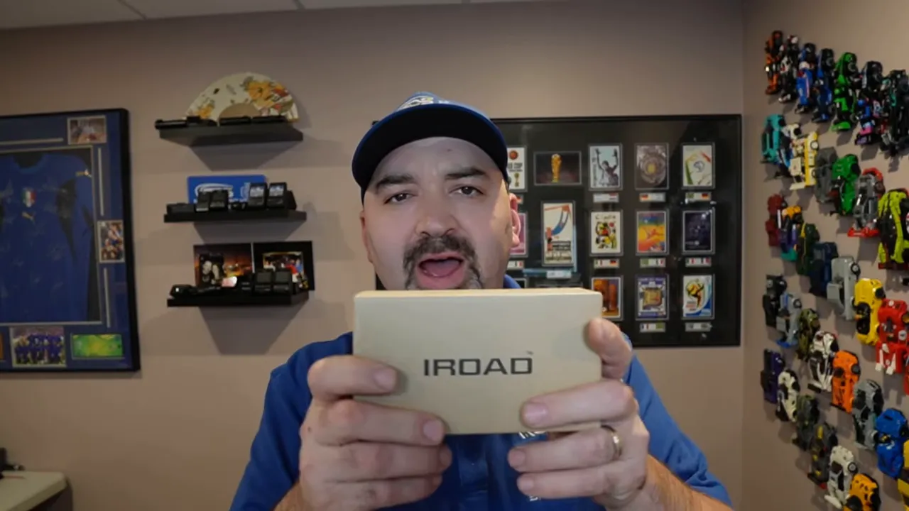 Presenter holding an IROAD product box in a demo room with dash cam samples on the wall