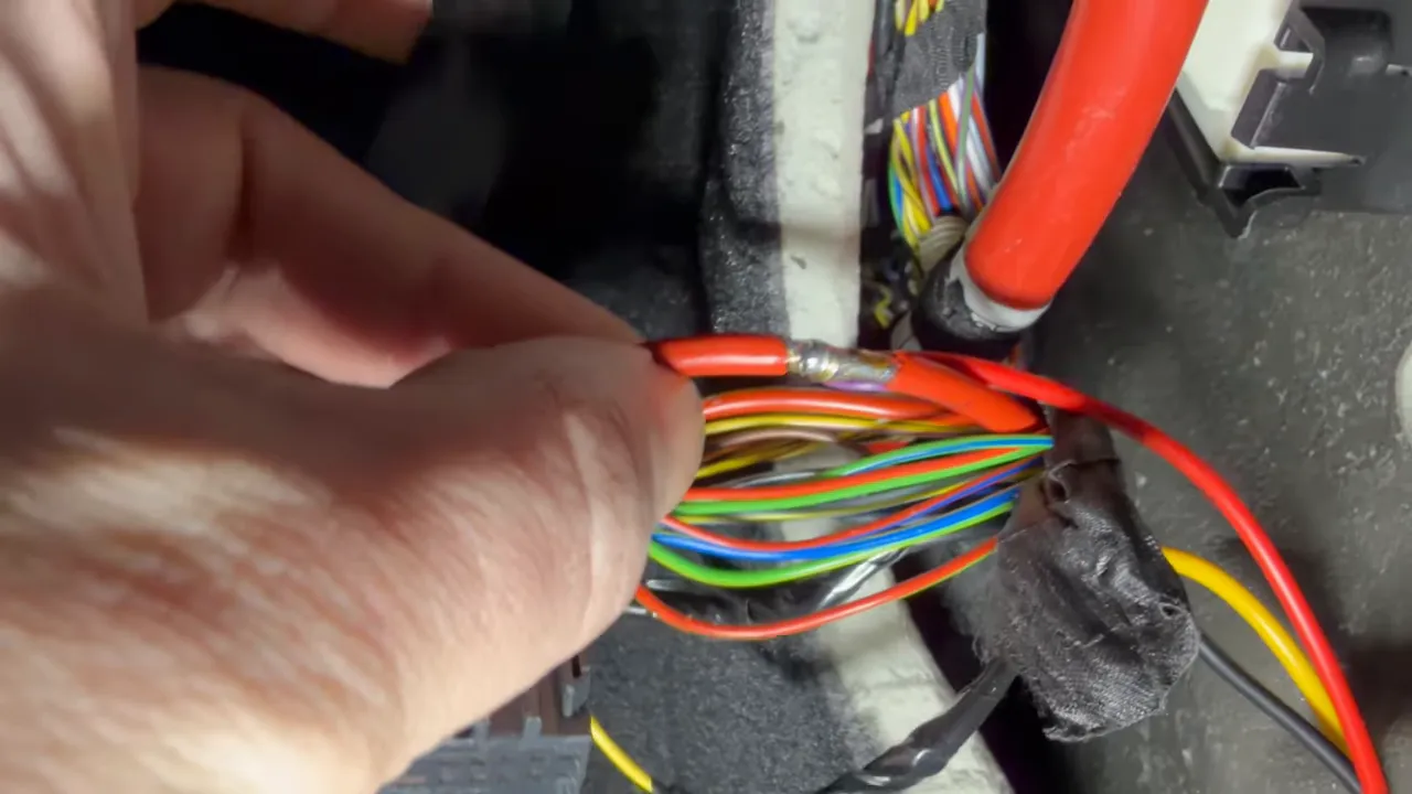 Hand holding red power wire showing a solder joint on a BMW wiring harness in the passenger kick panel.