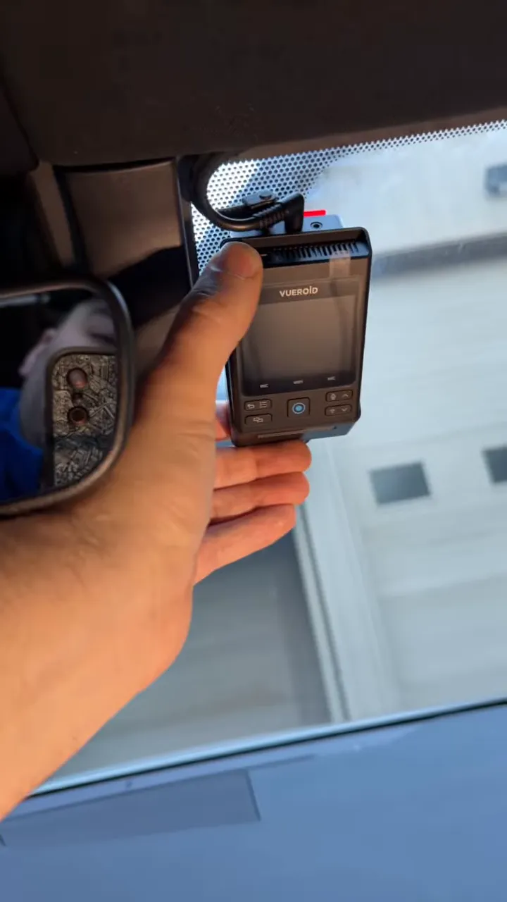 Hand holding a dash cam while its cable is routed into the headliner trim near the rearview mirror
