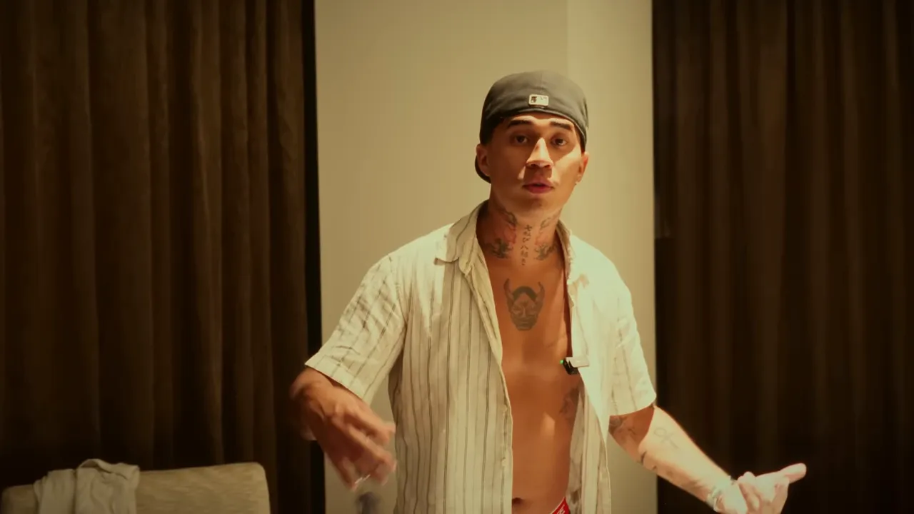 Homme en chemise ouverte et casquette parlant face caméra dans une chambre d'hôtel