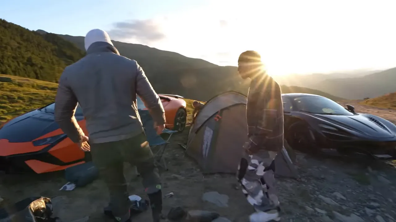 Campement en montagne au coucher du soleil avec tente, deux supercars et personnes en train d’installer le camp