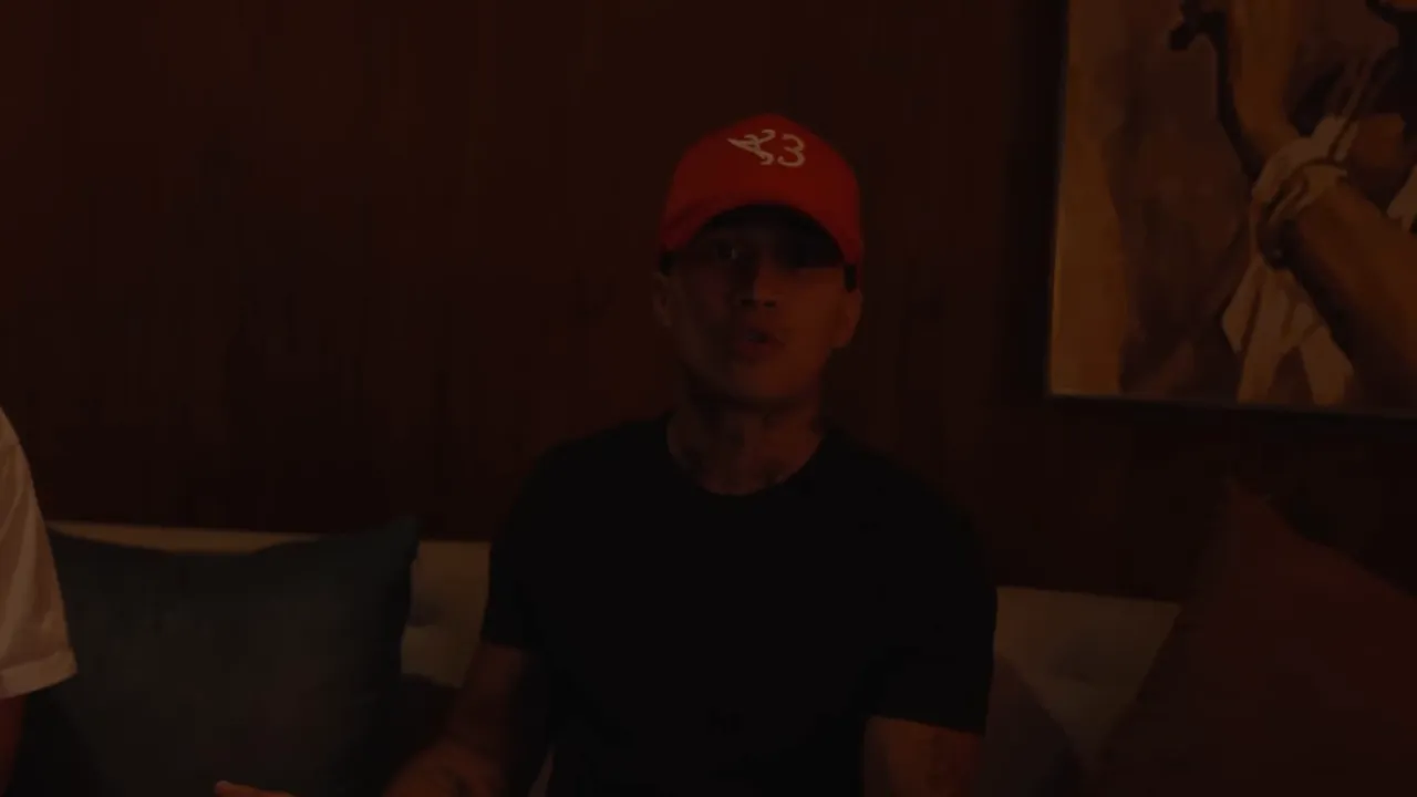 Personne assise en casquette rouge regardant la caméra et parlant dans un salon faiblement éclairé