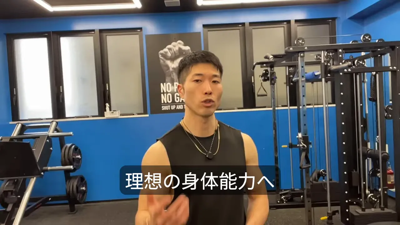 ジム背景でカメラに向かって話すトレーナーの中距離ショット（設備が見える）