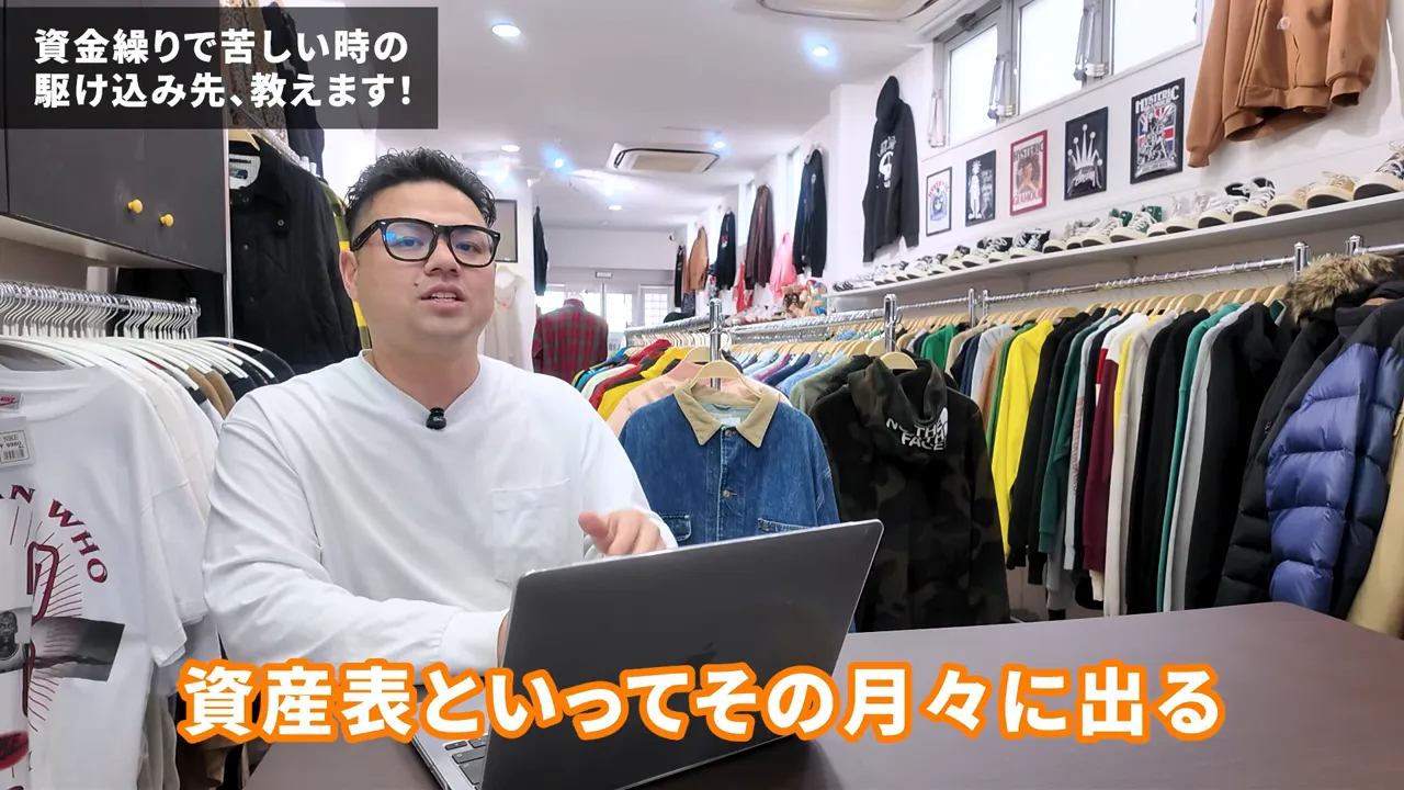 店内でノートパソコンを前に説明する男性、後ろに洋服が並ぶ明瞭な構図