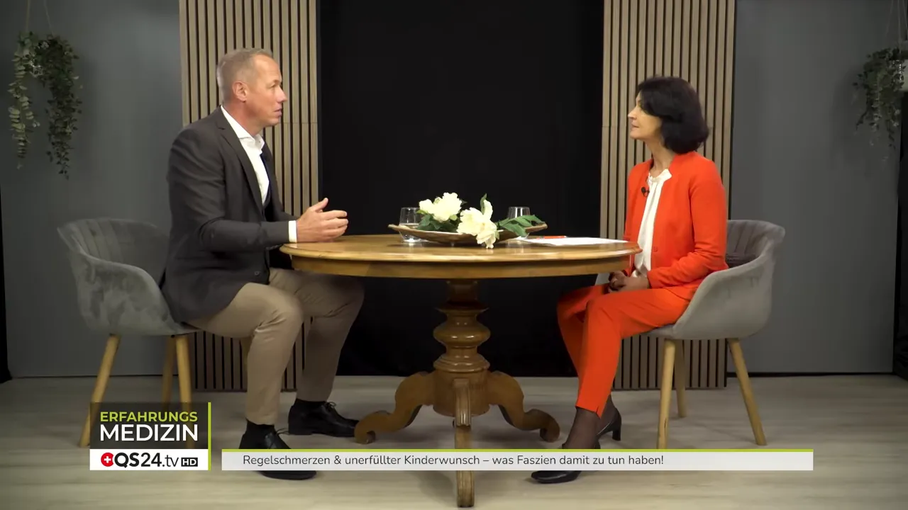 Zwei Personen sitzen sich gegenüber an einem runden Tisch im Studio, Beine und Haltung deutlich sichtbar.