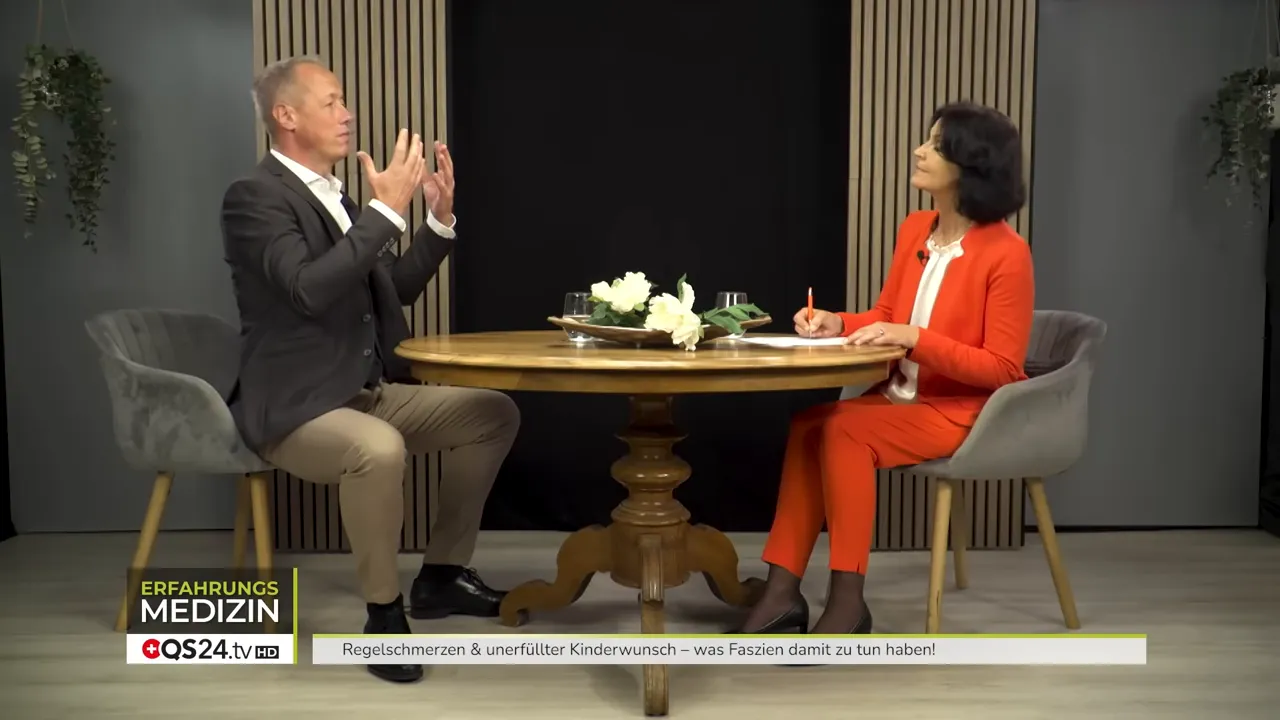 Breiter Bildausschnitt: David Boeger und Moderatorin am Tisch, er erklärt gestikulierend