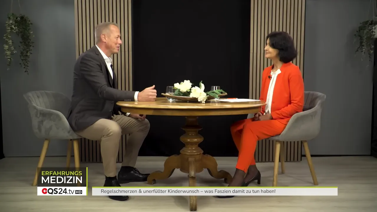 Zwei Gesprächspartner sitzen sich gegenüber an einem runden Holztisch im Studio; klare, scharfe Gesamtaufnahme ohne störende Einblendungen.