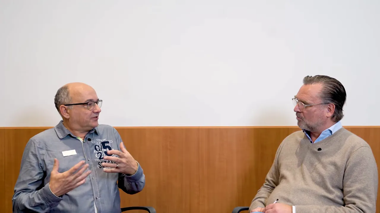 Zwei Experten im Gespräch vor neutraler Wand, einer erklärt gestikulierend.