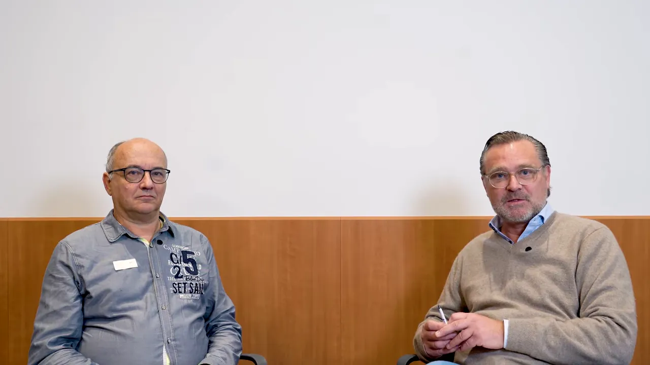 Zwei Männer frontal sitzend vor weißer Wand mit Holzpaneel