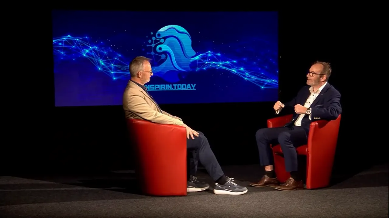 Zwei Gesprächspartner in roten Sesseln vor einem großen Bildschirm mit digitaler Grafik und Logo