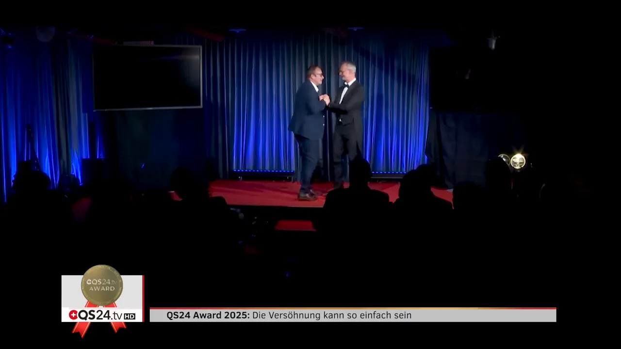QS24 AWARD 2025: Versöhnung in der Medizin — Preisträgerinnen und Preisträger 13 Zwei Personen auf der Bühne beim herzlichen Handschlag während einer Preisübergabe, blau beleuchteter Vorhang