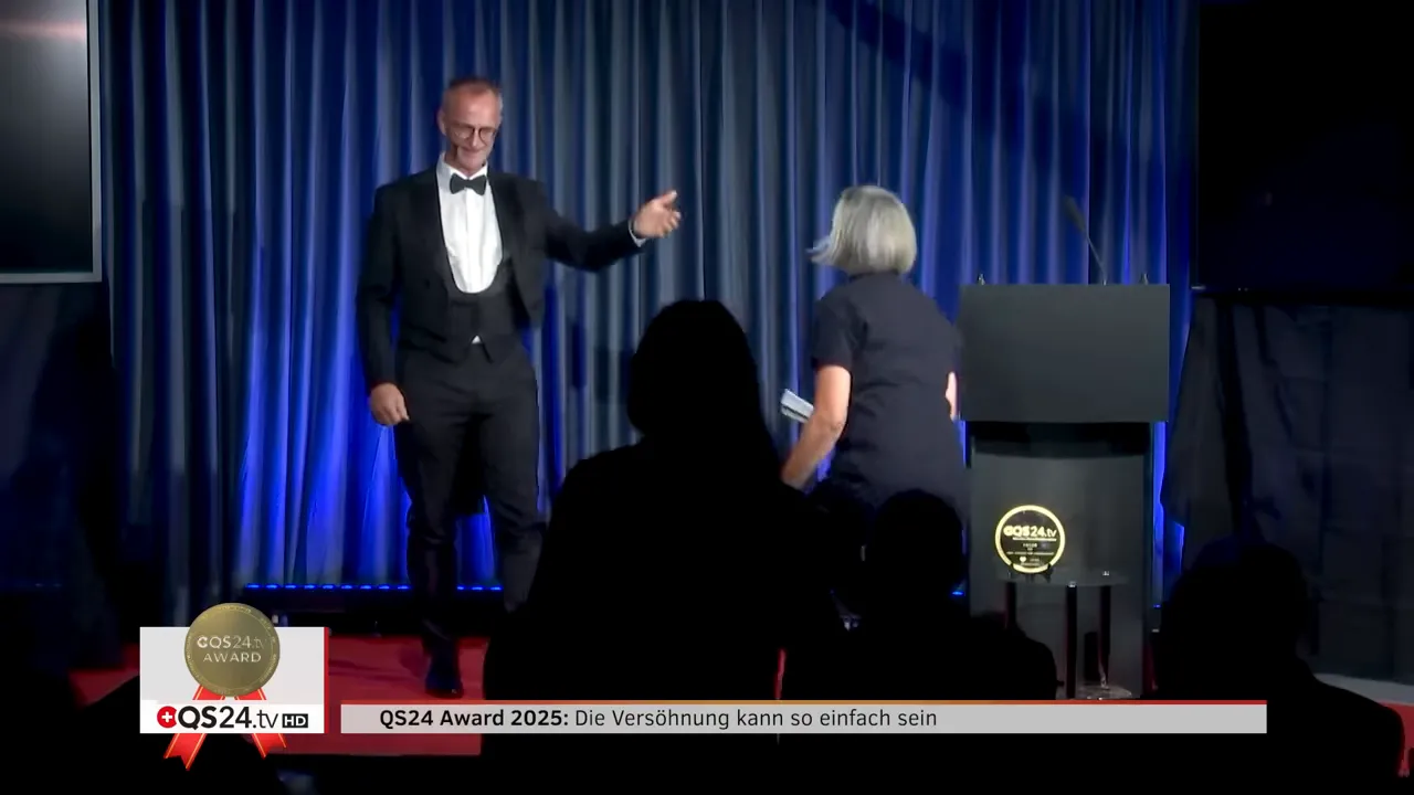 QS24 AWARD 2025: Versöhnung in der Medizin — Preisträgerinnen und Preisträger 11 Moderator in Smoking begrüsst Preisträgerin beim Betreten der Bühne vor blauem Vorhang