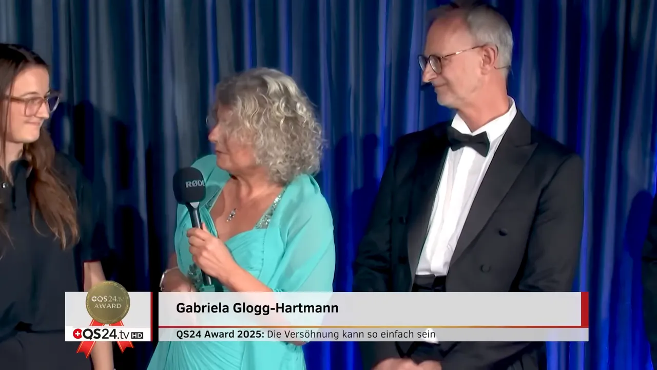 QS24 AWARD 2025: Versöhnung in der Medizin — Preisträgerinnen und Preisträger 14 Drei Personen auf der Bühne, Gabriela Glogg‑Hartmann in der Mitte, blauer Vorhang im Hintergrund