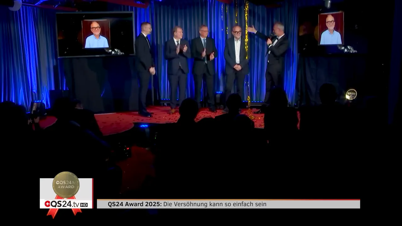 Moderator deutet auf den Preisträger auf der Bühne; zwei Monitore im Hintergrund zeigen ein Porträt des Gewinners