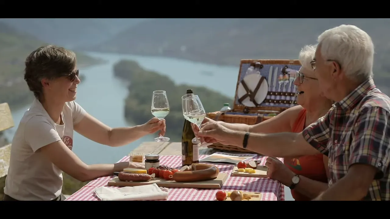 Mehrere Personen bei einem Picknick stoßen mit Weingläsern an vor einer Flusslandschaft
