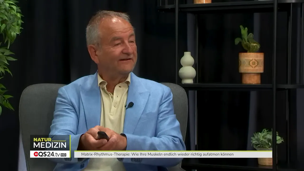 Wie Ihre Muskeln wieder aufatmen können - Interview mit Dr. Ulrich Randoll, Begründer der Matrix‑Rhythmus‑Therapie 7 Interviewgast hält ein Steuergerät/Remote und zeigt die Bedienung des Therapiegeräts im Studiokontext.