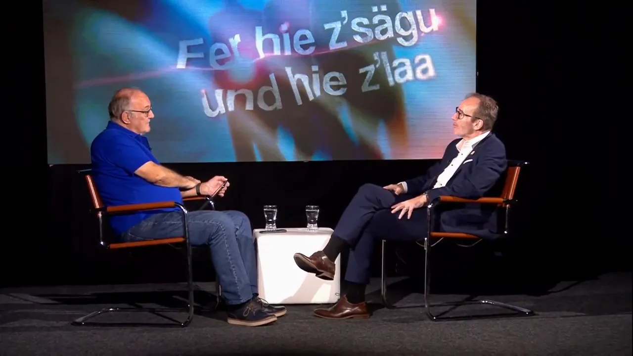 Zwei Männer sitzen sich in Sesseln gegenüber auf einer Bühne mit großer Leinwand im Hintergrund