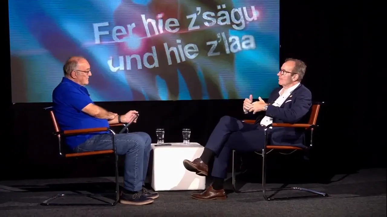 Zwei Personen im Studiogespräch auf Stühlen mit Tisch und Wassergläsern, Bildschirm im Hintergrund.