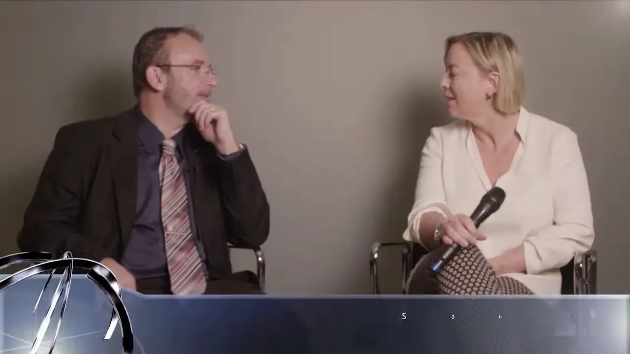 Zwei Personen in einem TV-Interview, eine hält ein Mikrofon, grafische Einblendung am unteren Bildrand